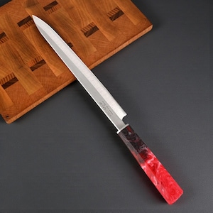 May include: A long, stainless steel knife with a red and black marbled handle rests on a wooden cutting board. The blade is sharp and pointed, and the handle is a vibrant mix of red and black. The knife is designed for slicing and cutting.