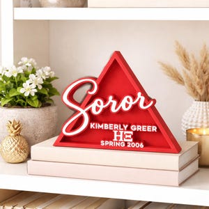 Puede incluir: Un letrero triangular rojo de madera con el texto en cursiva blanca Soror, el nombre Kimberly Greer, las letras griegas HΞ y el texto Spring 2006. El letrero se exhibe sobre dos libros apilados, mostrando un diseño audaz y dimensional.