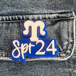 May include: Blue and white enamel pin with the text "Spr 24" and a white stylized letter "M" above the text.