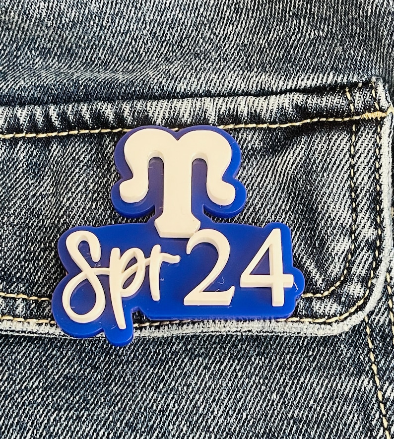 May include: Blue and white enamel pin with the text "Spr 24" and a white stylized letter "M" above the text.