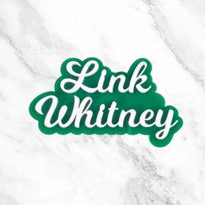 May include: A green and white decorative sign with the words "Link Whitney" in a cursive font. The text is layered on a green cloud-shaped background. The sign is set against a white marble background.