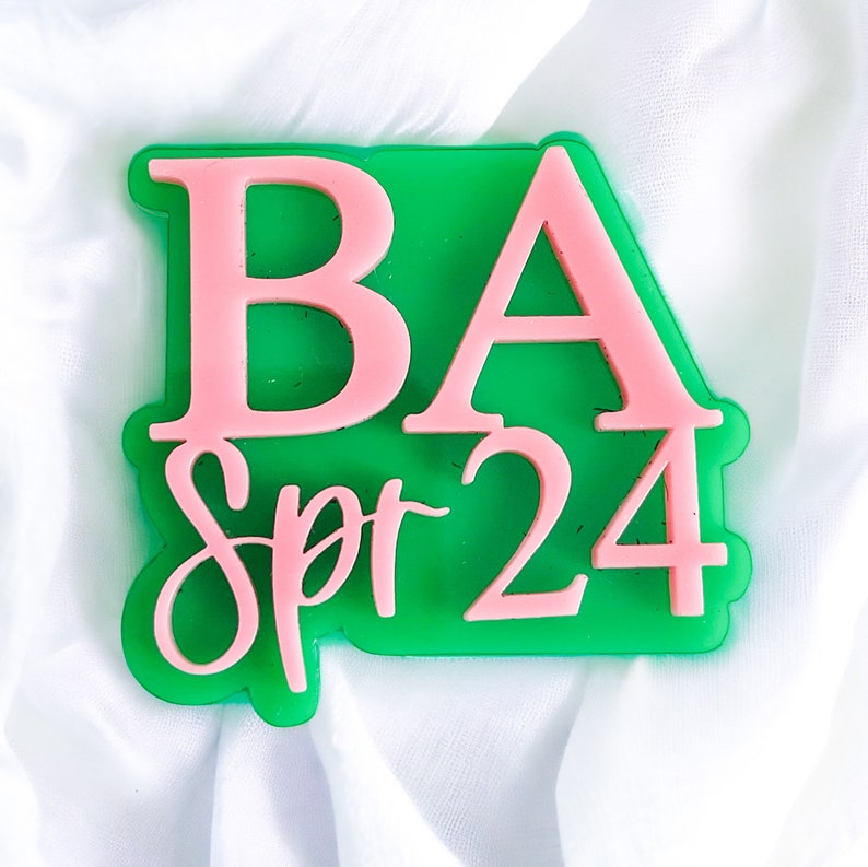 May include: Green and pink plastic sign with the text "BA Spr 24" in a cursive font.