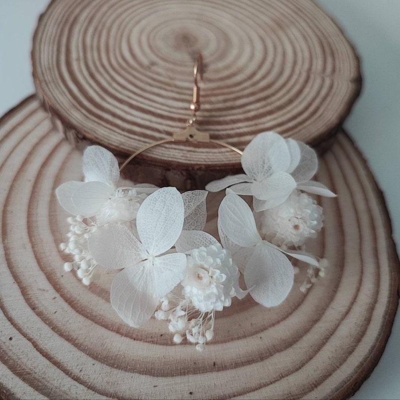 Op de afbeelding: Een paar gouden oorringen versierd met delicate witte gedroogde bloemen. De oorbellen hebben een cluster van kleine, getextureerde bloemen en grotere, bloembladachtige bloemen, wat een natuurlijk en elegant ontwerp cre&euml;ert. Gepresenteerd op een houten schijf.