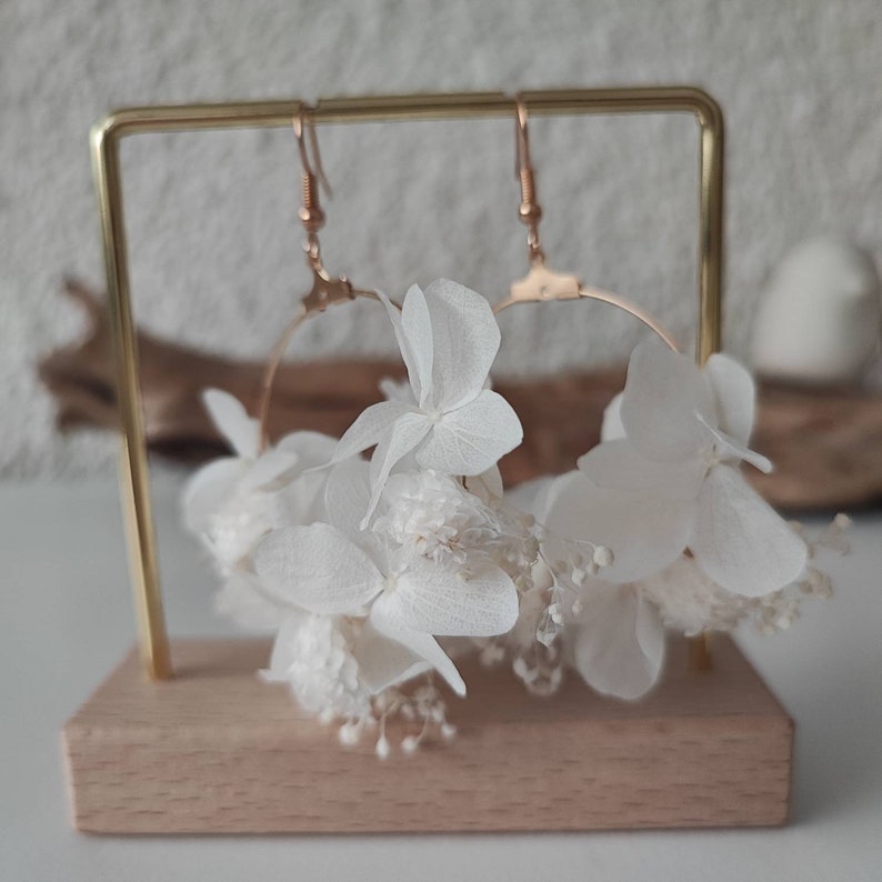 Op de afbeelding: Gouden oorringen met witte gedroogde bloemen, tentoongesteld op een gouden standaard met een houten voet. De oorbellen tonen een delicaat bloemendesign en voegen een vleugje natuurlijke elegantie toe aan de sieraden.