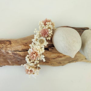 Op de afbeelding: Een bloemen haaraccessoire met gedroogde bloemen in crème- en roze tinten. De bloemen zijn gerangschikt op een flexibele basis, geschikt voor diverse kapsels. Het accessoire wordt gepresenteerd op een stuk drijfhout.