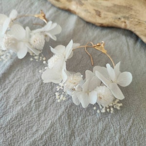 Op de afbeelding: Een paar goudkleurige oorringen versierd met witte gedroogde bloemen. De oorbellen hebben een delicate opstelling van kleine, getextureerde bloemen en grotere, bloemblad-vormige bloemen, waardoor een natuurlijk, elegant ontwerp ontstaat.