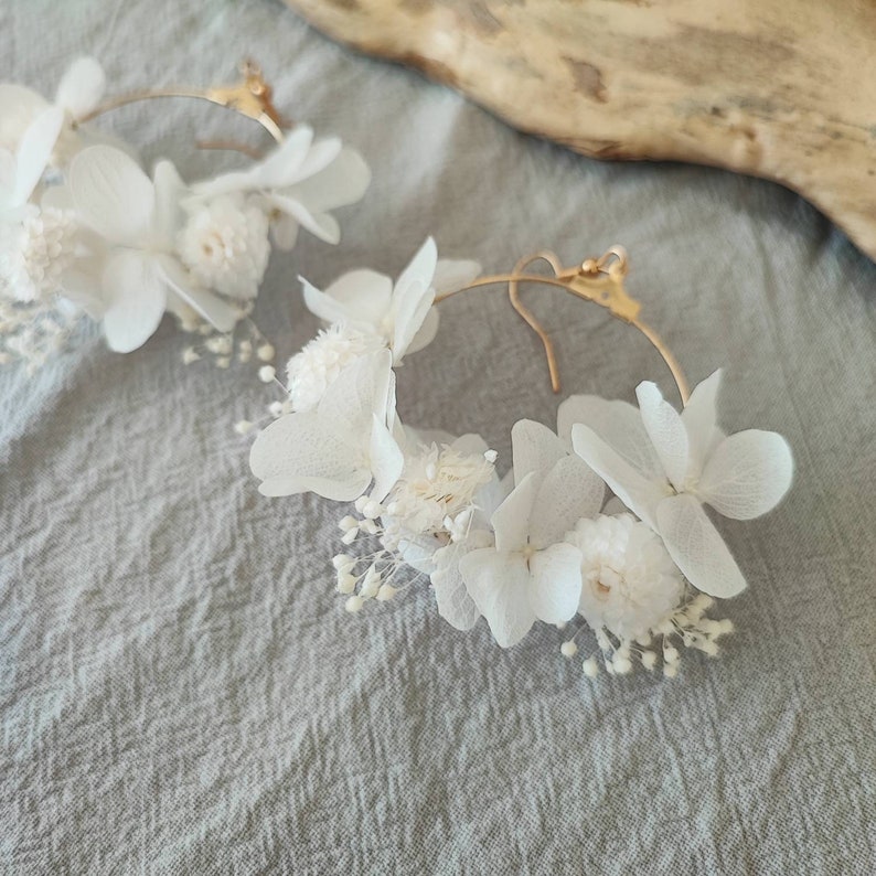 Op de afbeelding: Een paar goudkleurige oorringen versierd met witte gedroogde bloemen. De oorbellen hebben een delicate opstelling van kleine, getextureerde bloemen en grotere, bloemblad-vormige bloemen, waardoor een natuurlijk, elegant ontwerp ontstaat.