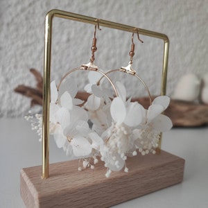 Op de afbeelding: Gouden oorringen met witte gedroogde bloemen. De oorbellen worden gepresenteerd op een gouden standaard met een houten basis. Het bloemstuk bevat kleine, delicate witte bloesems.