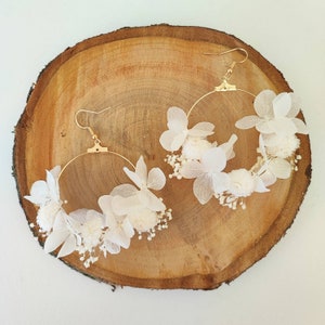 Op de afbeelding: Een paar goudkleurige oorringen versierd met delicate witte bloemen en kleine witte bloemaccenten. De oorbellen worden gepresenteerd op een natuurlijk houten oppervlak, wat hun elegante ontwerp en bloementhema benadrukt.