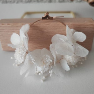 Op de afbeelding: Een delicate, witte bloemen oorring. De oorring heeft een goudkleurige metalen ring versierd met clusters van witte gedroogde bloemen, waaronder hortensia's en kleinere, getextureerde bloemen. De oorring wordt gepresenteerd op een licht houten blok.