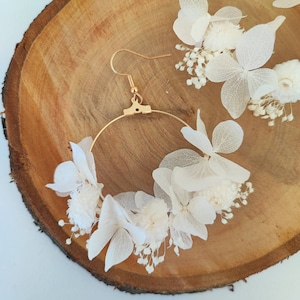 Op de afbeelding: Goudkleurige oorringen versierd met delicate witte bloemen. De oorbellen hebben een cirkelvormig ontwerp met een haaksluiting. Extra bloemaccenten zijn in de buurt verspreid, alles tegen een houten achtergrond.