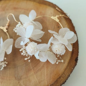 Op de afbeelding: Een paar goudkleurige oorringen versierd met delicate witte bloemen en kleine, bolvormige details. De oorbellen worden gepresenteerd tegen een achtergrond van natuurlijke houtnerf, wat het ingewikkelde bloemendesign benadrukt.