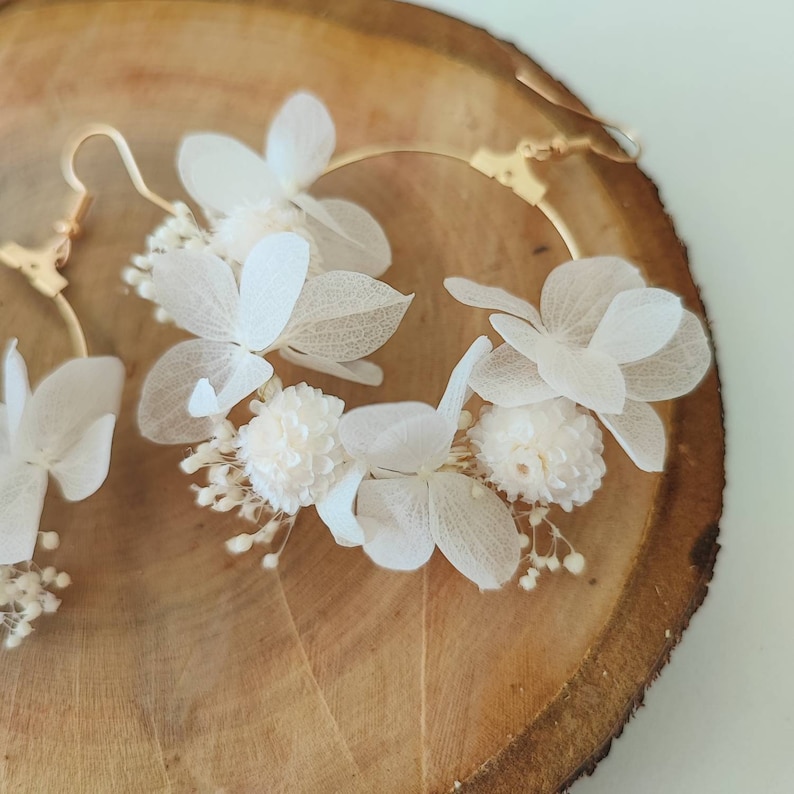 Op de afbeelding: Een paar goudkleurige oorringen versierd met delicate witte bloemen en kleine, bolvormige details. De oorbellen worden gepresenteerd tegen een achtergrond van natuurlijke houtnerf, wat het ingewikkelde bloemendesign benadrukt.