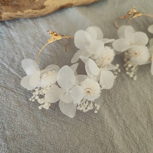 Op de afbeelding: Een paar goudkleurige oorringen versierd met delicate witte gedroogde bloemen. De oorbellen hebben kleine, ronde bloemen en grotere, bloembladachtige bloemen, waardoor een natuurlijk, elegant ontwerp ontstaat. De oorbellen staan tegen een zachte grijze achtergrond.