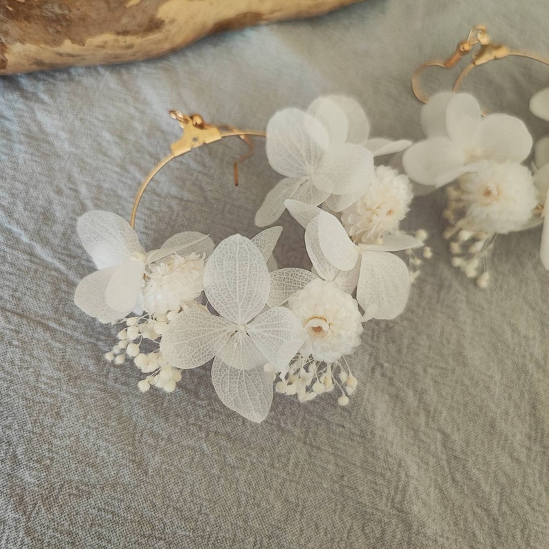 Op de afbeelding: Een paar goudkleurige oorringen versierd met delicate witte gedroogde bloemen. De oorbellen hebben kleine, ronde bloemen en grotere, bloembladachtige bloemen, waardoor een natuurlijk, elegant ontwerp ontstaat. De oorbellen staan tegen een zachte grijze achtergrond.
