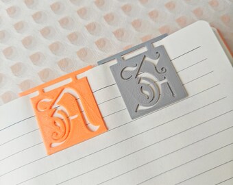 Op de afbeelding: Twee 3D-geprinte bladwijzerontwerpen, een oranje en een grijs, met een decoratieve letter 'A' en 'J' op elk.