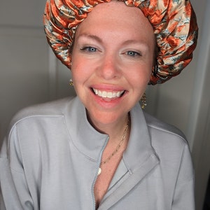 K&ouml;nnte beinhalten: Eine Frau mit einem strahlenden L&auml;cheln tr&auml;gt einen hellblauen Pullover mit Rei&szlig;verschluss und eine goldene Kettenkette. Sie tr&auml;gt auch eine Satin-Schlafhaube mit einem floralen Muster in Orange-, Braun- und Wei&szlig;t&ouml;nen.