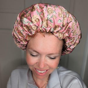 K&ouml;nnte beinhalten: Eine rosa Satin-Schlafhaube mit Blumenmuster. Die Haube ist oben gerafft und hat einen Kordelzug.