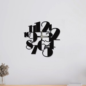 May include: A modern black wall clock with a minimalist design. The clock features cut-out numbers in a unique font, with the hands visible in the center. The clock is mounted on a white wall, above a wooden cabinet.