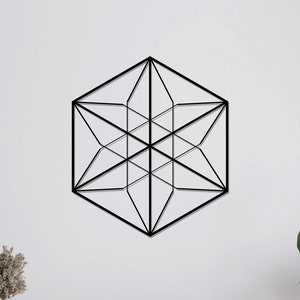 May include: Black geometric wall decor in the shape of a hexagon with a starburst design. The decor is hanging on a white wall above a wooden shelf with a plant on the right and a potted plant on the left.