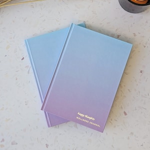 May include: Two blue and purple gradient journals with the text "happy thoughts WELLNESS JOURNAL" on the front.