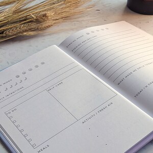 May include: A white journal with a purple cover. The journal has sections for daily tasks, self-care, meals, activities, and evening reflections. The journal is open to a page with a section for "What made me smile today?" and "Today I'm grateful for..."
