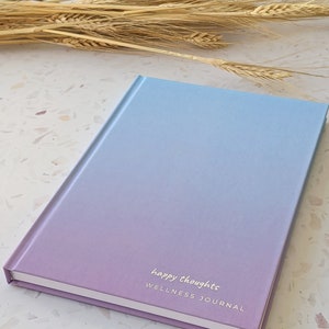 May include: A purple and blue ombre wellness journal with the text "happy thoughts" and "WELLNESS JOURNAL" on the front cover.