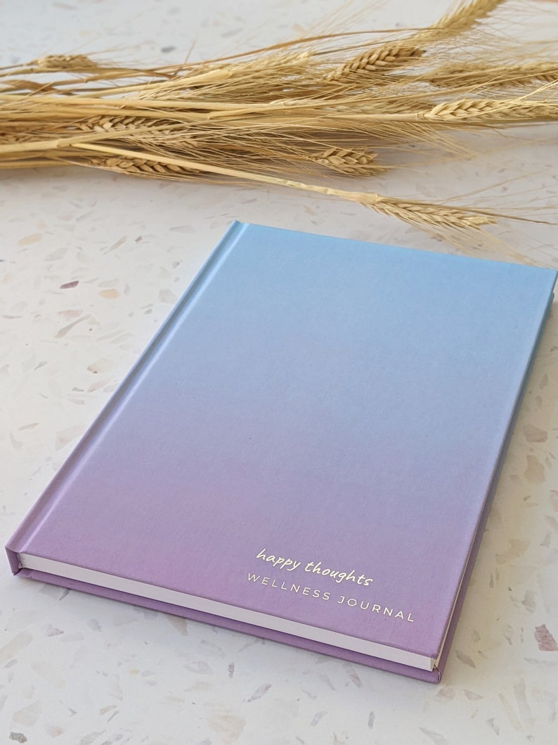 May include: A purple and blue ombre wellness journal with the text "happy thoughts" and "WELLNESS JOURNAL" on the front cover.