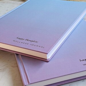 May include: Two pastel-colored journals stacked on top of each other. The top journal is light blue and purple with the text "happy thoughts WELLNESS JOURNAL" printed on the front. The bottom journal is a darker shade of purple.