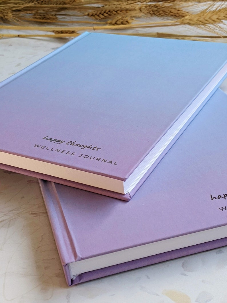 May include: Two pastel-colored journals stacked on top of each other. The top journal is light blue and purple with the text "happy thoughts WELLNESS JOURNAL" printed on the front. The bottom journal is a darker shade of purple.
