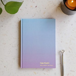 May include: A blue and purple ombre wellness journal with the text "happy thoughts" and "WELLNESS JOURNAL" on the front cover. A silver pen with a diamond-shaped top is next to the journal.