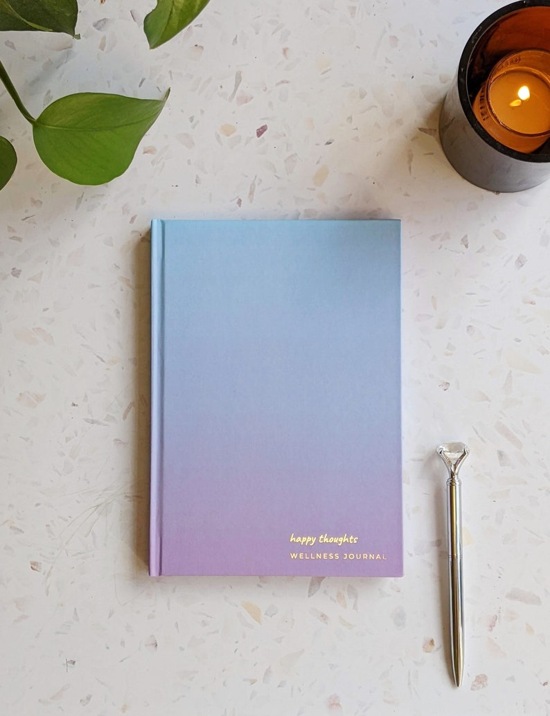 May include: A blue and purple ombre wellness journal with the text "happy thoughts" and "WELLNESS JOURNAL" on the front cover. A silver pen with a diamond-shaped top is next to the journal.