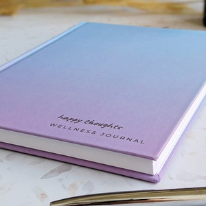 May include: A purple and blue ombre journal with the text "happy thoughts WELLNESS JOURNAL" on the cover.