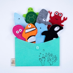 Peut inclure: Un ensemble de marionnettes en feutre d'animaux marins dans une enveloppe en feutre vert. Les marionnettes comprennent une tortue, une baleine, un dauphin, un poisson, un crabe et une pieuvre. L'enveloppe porte l'inscription "Little Bocco".