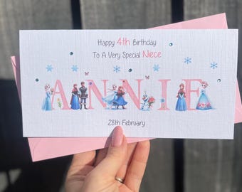 以下が含まれることがあります： ピンクと白のフローズンをテーマにした誕生日カード。カードには「Happy 4th Birthday To A Very Special Niece」と「ANNIE」が書かれており、フローズンのキャラクターが文字の中に描かれています。カードの下部には「28th February」と日付が書かれています。