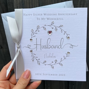 Op de afbeelding: Een zilveren huwelijksjubileumkaart met een witte achtergrond en zilveren tekst. De kaart luidt: "Happy Silver Wedding Anniversary To My Wonderful Husband Nicholas 10 september 2023". Een zilveren lint is om de kaart gebonden.