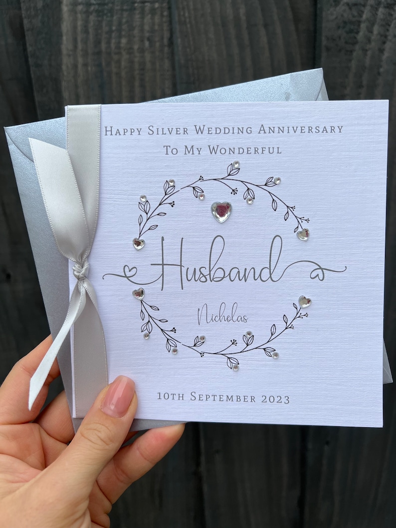 Op de afbeelding: Een zilveren huwelijksjubileumkaart met een witte achtergrond en zilveren tekst. De kaart luidt: "Happy Silver Wedding Anniversary To My Wonderful Husband Nicholas 10 september 2023". Een zilveren lint is om de kaart gebonden.