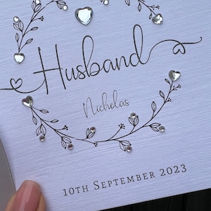 Op de afbeelding: Een witte kaart met het woord "Husband" in elegant schrift, omcirkeld door een bloemenkrans en versierd met hartvormige edelstenen. De naam "Nicholas" staat eronder, en de datum "10th September 2023" is onderaan gedrukt.