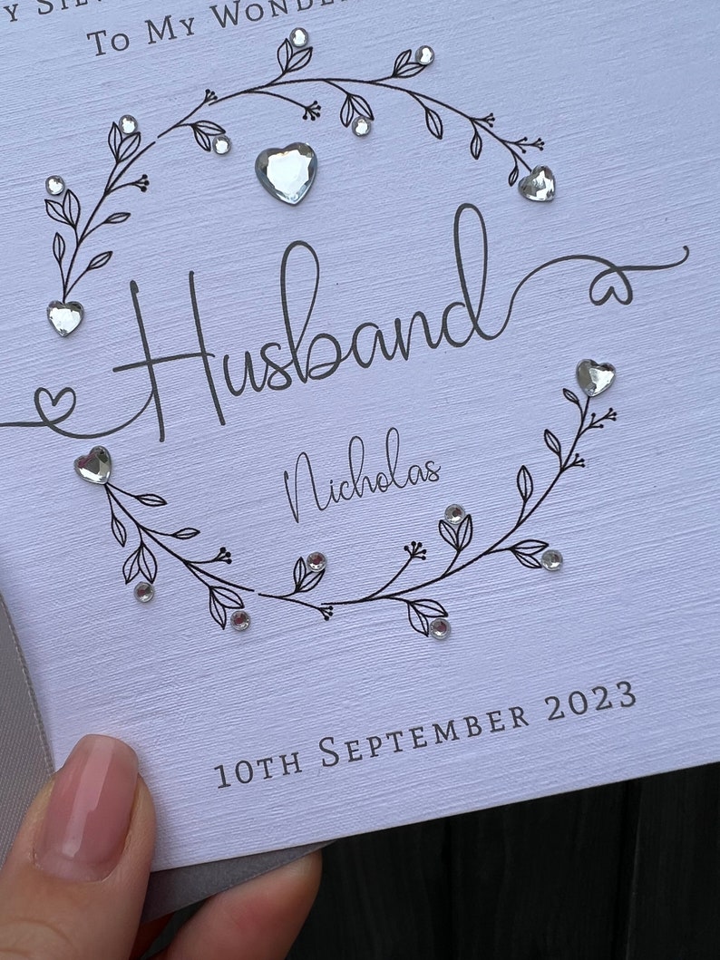 Op de afbeelding: Een witte kaart met het woord "Husband" in elegant schrift, omcirkeld door een bloemenkrans en versierd met hartvormige edelstenen. De naam "Nicholas" staat eronder, en de datum "10th September 2023" is onderaan gedrukt.