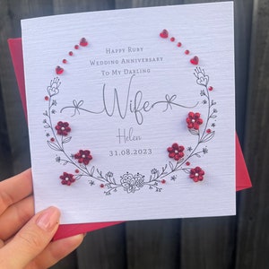 May include: A white card with a red floral wreath and red glitter hearts. The card says "Happy Ruby Wedding Anniversary To My Darling Wife Helen 31.08.2023".