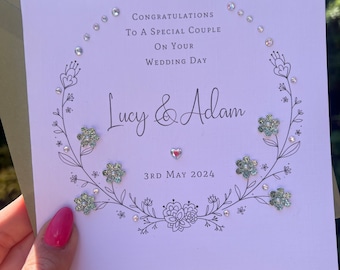 May include: A white card with a floral wreath design and the text "Congratulations to a special couple on your wedding day Lucy & Adam 3rd May 2024".