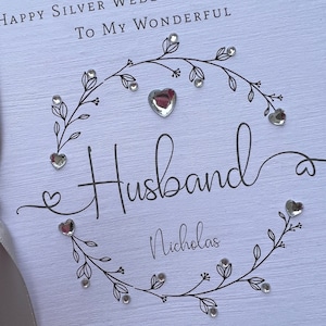 Op de afbeelding: Een witte kaart met de tekst "Happy Silver Wedding Anniversary To My Wonderful Husband Nicholas" en de datum "10 september 2023". De kaart is versierd met een bloemenontwerp en hartvormige versieringen.