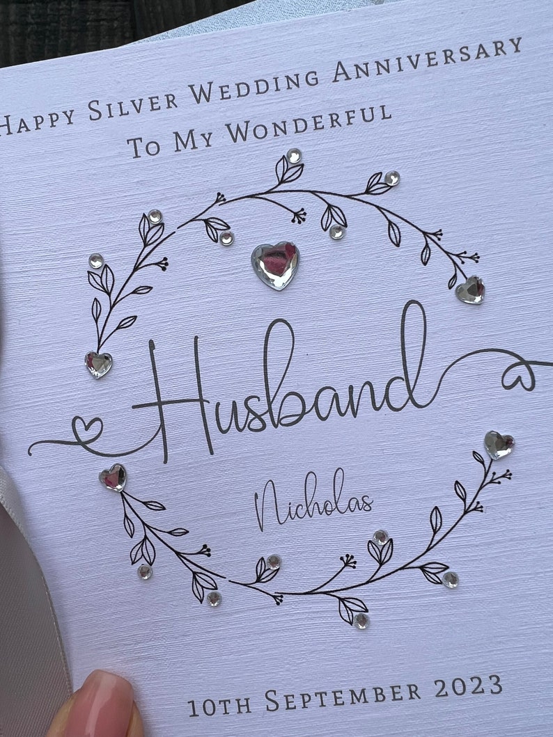 Op de afbeelding: Een witte kaart met de tekst "Happy Silver Wedding Anniversary To My Wonderful Husband Nicholas" en de datum "10 september 2023". De kaart is versierd met een bloemenontwerp en hartvormige versieringen.