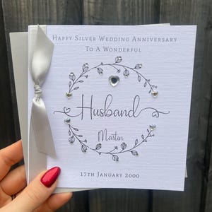 Op de afbeelding: Een zilveren huwelijksjubileumkaart met de tekst "Happy Silver Wedding Anniversary To A Wonderful Husband Martin 17th January 2000". De kaart is versierd met een bloemmotief en een zilveren lint.