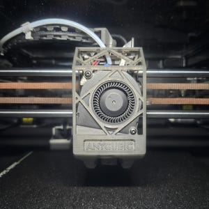 Puede incluir: Primer plano del cabezal de una impresora 3D con un ventilador y el logotipo "ANYCUBIC". El cabezal es gris y tiene un diseño geométrico. Las correas y otros componentes de la impresora son visibles en el fondo.