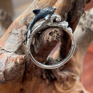 May include: A silver ring featuring a black and silver dolphin leaping over a wave. The ring is set against a natural wood background, highlighting the aquatic theme. The dolphin has a black body and silver accents.