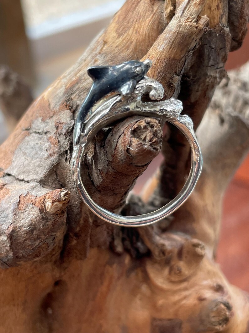 May include: A silver ring featuring a black and silver dolphin leaping over a wave. The ring is set against a natural wood background, highlighting the aquatic theme. The dolphin has a black body and silver accents.