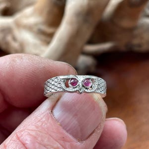 Könnte beinhalten: Ein silberner Ring mit einem Eulenmotiv. Der Ring hat zwei runde, rosa Edelsteine als Augen. Das Gesicht der Eule ist mit strukturierten Mustern detailliert. Der Ring wird vor einem verschwommenen Hintergrund gehalten.