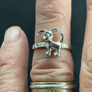 May include: A silver-toned ring featuring a small dog figure with floppy ears and a curled tail. The band of the ring is decorated with a repeating bone pattern. The ring is worn on a finger above a stack of two other metallic rings.
