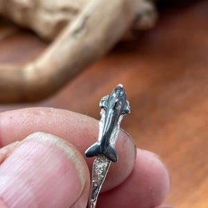 May include: A silver ring featuring a detailed dolphin design. The dolphin is depicted with a sleek, curved body and a prominent dorsal fin. The ring's band has a textured, wave-like pattern, adding to the aquatic theme.